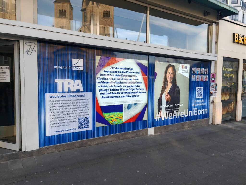 Schaufensterfront der Universität Bonn mit großem Plakat zu TRA (Transdisziplinäre Forschungsbereiche), erklärendem Text, Fotos von Jacqueline Lorenzen, einem Zitat zur Klimaanpassung sowie verschiedenen QR-Codes und dem Slogan #WeAreUniBonn.