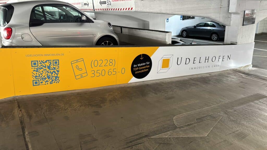 Werbetafel der Firma Udelhofen Immobilien GmbH in einem Parkhaus mit gelbem und weißem Hintergrund, QR-Code, Telefonnummer und Slogan "Ihr Makler für TOP-Gewerbeimmobilien", daneben parkende Autos.