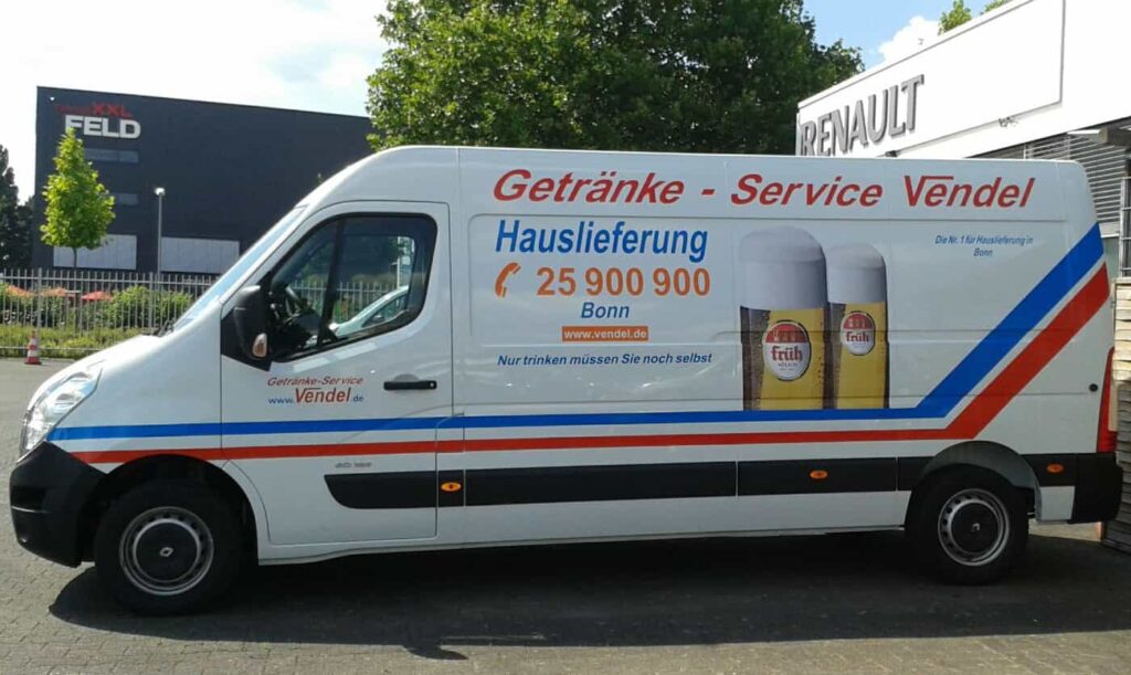 Weißer Lieferwagen mit Werbung für "Getränke-Service Vendel", inklusive Kontaktinformationen, Slogan "Hauslieferung", zwei Gläser Kölsch und bunten Streifen in Blau und Rot; geparkt vor Gebäuden mit den Schildern "FELD" und "RENAULT".