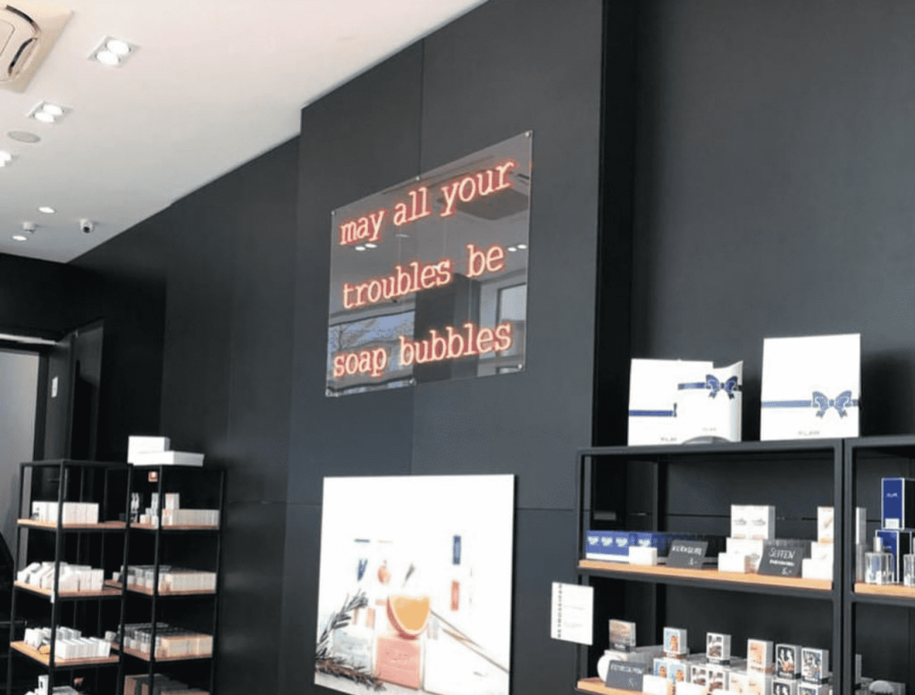 Innenansicht eines modernen Geschäfts mit schwarzen Wänden und Regalen mit Kosmetikprodukten; an der Wand hängt ein Leuchtschild mit der Aufschrift „may all your troubles be soap bubbles“.