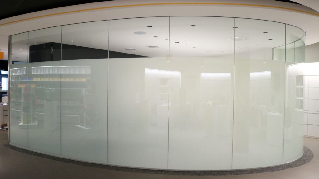 Rundes Zimmer mit einer Glaswand, deren unterer Bereich milchig-mattiert ist; dahinter schemenhaft Regale und Lichter sichtbar, modernes und lichtes Innendesign.