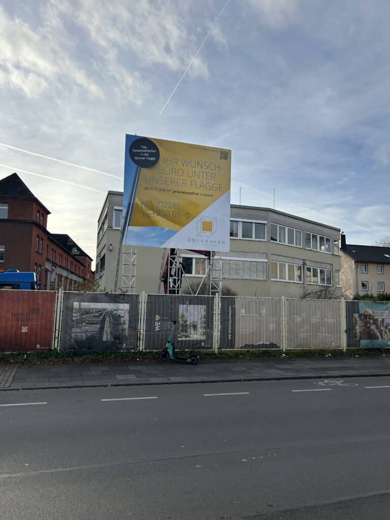 Großes Werbeplakat mit dem Text 'Ihr Wunsch-Büro unter unserer Flagge' und weiteren Informationen zu mietbaren Gewerbeflächen steht vor einem Bürogebäude, im Vordergrund ein Elektro-Scooter auf dem Gehweg.