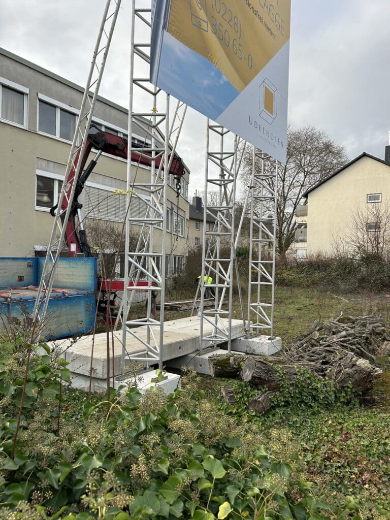 Großes Werbeschild auf einem Metallgerüst mit Betonfundament, im Vordergrund ein Haufen Baumstämme und Efeupflanzen auf einer unbebauten Grünfläche; dahinter ein Kranfahrzeug und mehrere Wohngebäude.