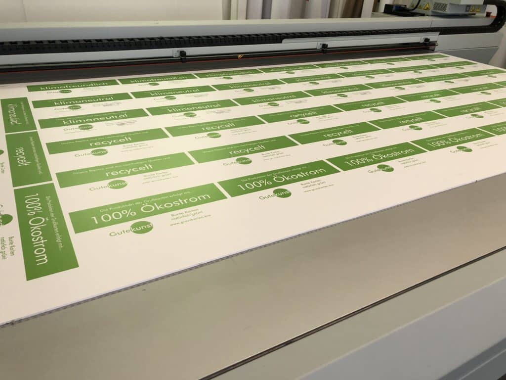 Großer Druckbogen mit mehrfach aufgedruckten, grünen Etiketten mit verschiedenen Umwelt- und Nachhaltigkeitssiegeln wie "klimaneutral", "recycelt" und "100% Ökostrom" auf weißem Untergrund in einer Druckerei.