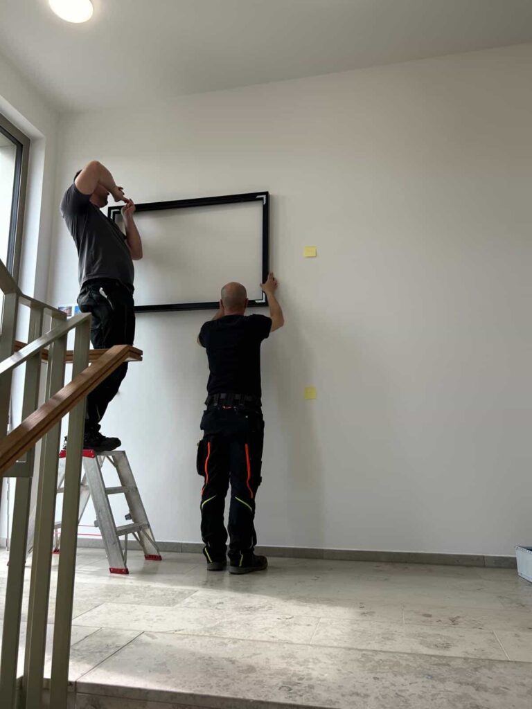 Zwei Handwerker montieren einen rechteckigen schwarzen Rahmen an eine weiße Wand; einer steht auf einer Leiter, der andere steht auf dem Boden und beide arbeiten konzentriert.