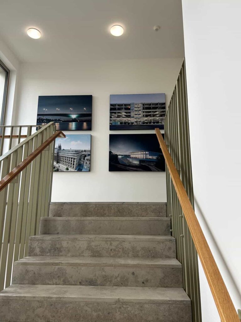 Betontreppe in einem modernen Gebäude mit grünen Geländern und Holzhandläufen, an der weißen Wand hängen vier gerahmte Fotografien von Gebäuden und Stadtansichten.