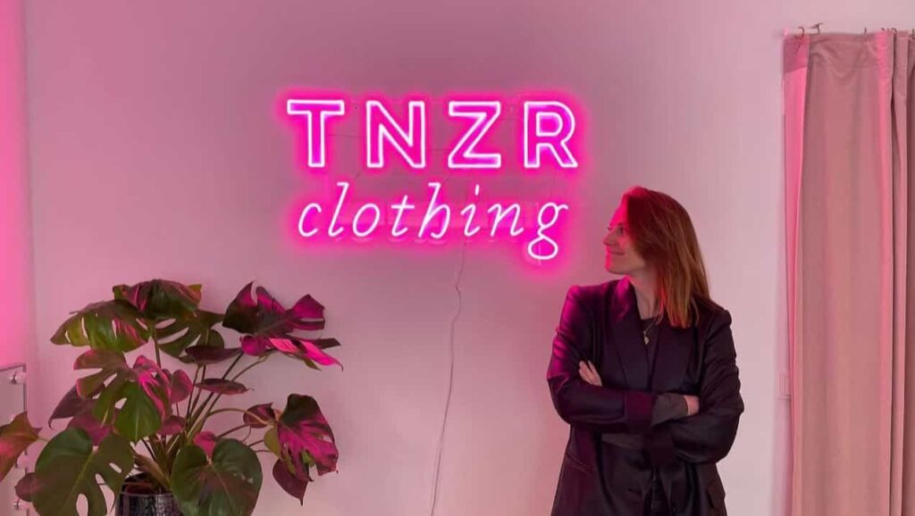 Frau in dunkler Kleidung steht vor einer weißen Wand mit neonpinker Leuchtschrift 'TNZR clothing', links daneben eine große grüne Zimmerpflanze.