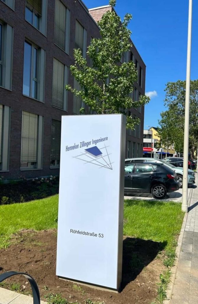 Helles Firmenschild mit der Aufschrift "Henneker Zillinger Ingenieure" und "Röhfeldstraße 53" vor einem modernen Bürogebäude, im Hintergrund sieht man Autos, einen Baum und einen blauen Himmel.