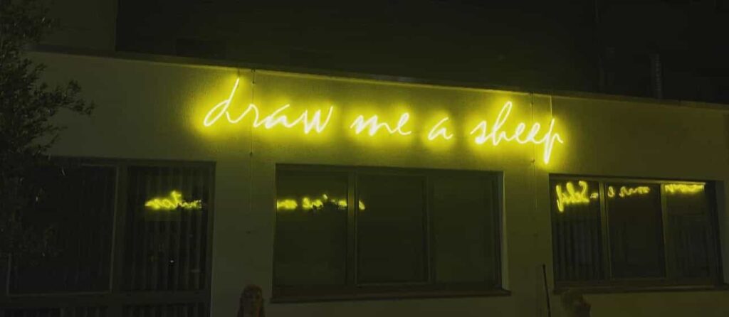 Leuchtschrift in gelbem Neon mit dem Text „draw me a sheep“ an einer Hauswand bei Nacht.