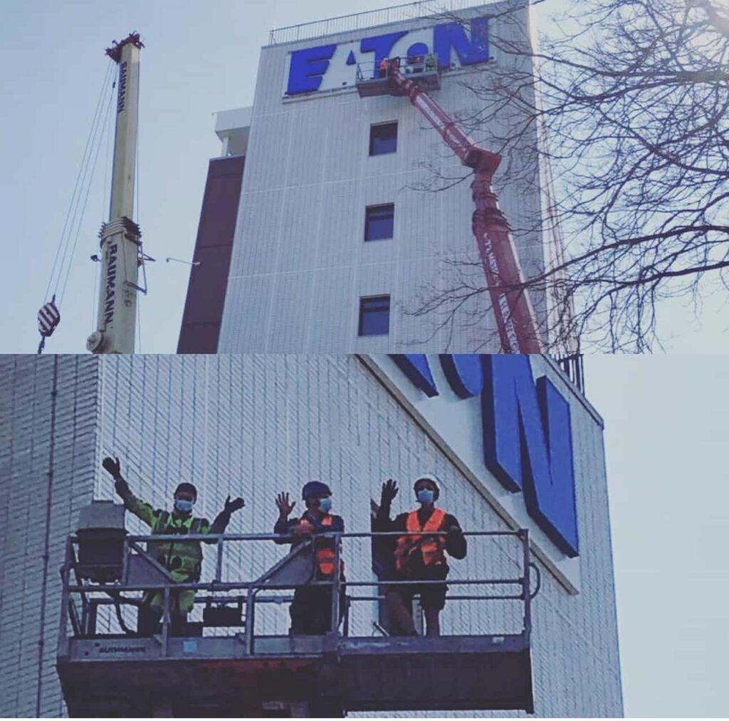 Zwei Fotos: Oben ein Kran und eine Hebebühne, auf der Arbeiter an einem hohen Gebäude das große blaue Firmenlogo "EATON" montieren; unten drei Bauarbeiter in Schutzkleidung und mit Masken winken von der Hebebühne in die Kamera.