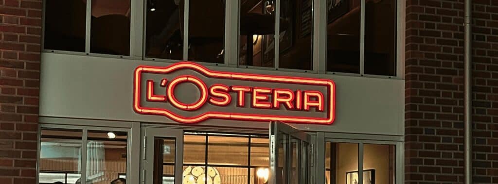 Leuchtendes Neonschild mit dem Schriftzug „L’Osteria“ an der Fassade eines Restaurants über dem Eingang.