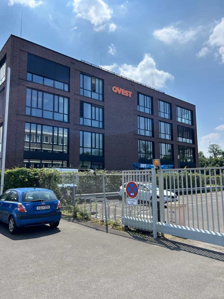 Modernes Bürogebäude mit großem Ovest-Schriftzug an der Fassade, großem Glasfenstern und davor ein Parkplatz mit Zaun, einem blauen Auto und einem Verkehrsschild, unter blauem Himmel mit Wolken.
