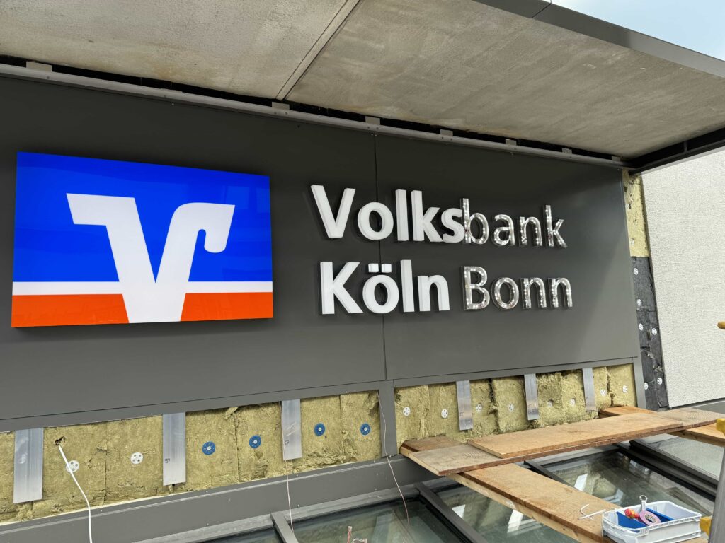 Bauschild mit dem Logo der Volksbank und der Aufschrift „Volksbank Köln Bonn“ an einer Fassade, bei der Bauarbeiten offensichtlich noch im Gange sind.