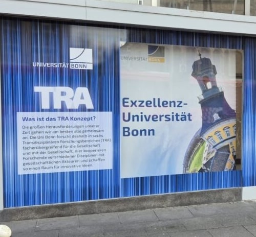 Werbeplakat der Universität Bonn mit dem Text 'TRA' und 'Exzellenz-Universität Bonn', daneben eine kurze Erklärung zum TRA-Konzept und ein Bild eines Kirchturms mit blauen und gelben Farbakzenten.