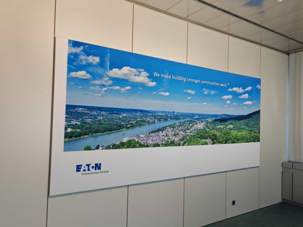 Großes Wandbild in einem Büro zeigt eine Panoramaaufnahme einer Stadt an einem Fluss mit bewaldeten Hügeln, blauem Himmel und Wolken; oben steht der Slogan "We make building stronger communities work" und unten das Logo von EATON.